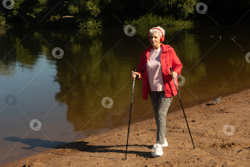 Скачать Седовласая женщина прогуливается с трекинговыми палками по пляжу возле озера фотосток Ozero