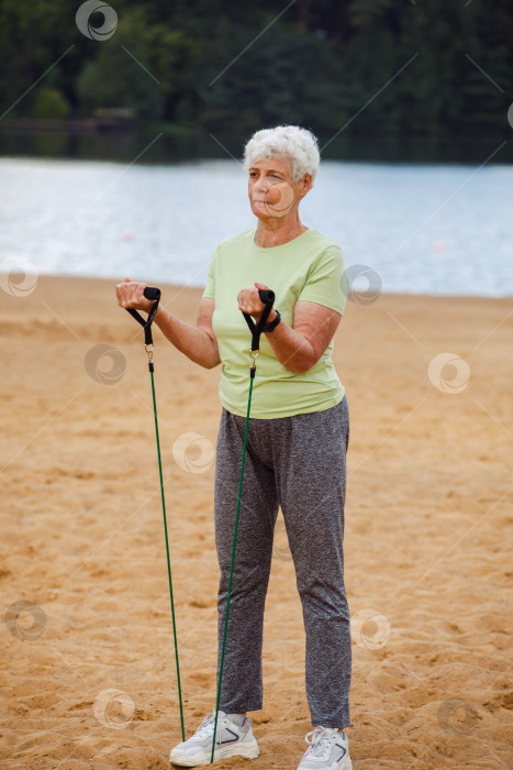 Скачать Пожилая женщина в спортивной одежде утром делает зарядку на открытом воздухе на пляже, используя эластичные резинки. фотосток Ozero