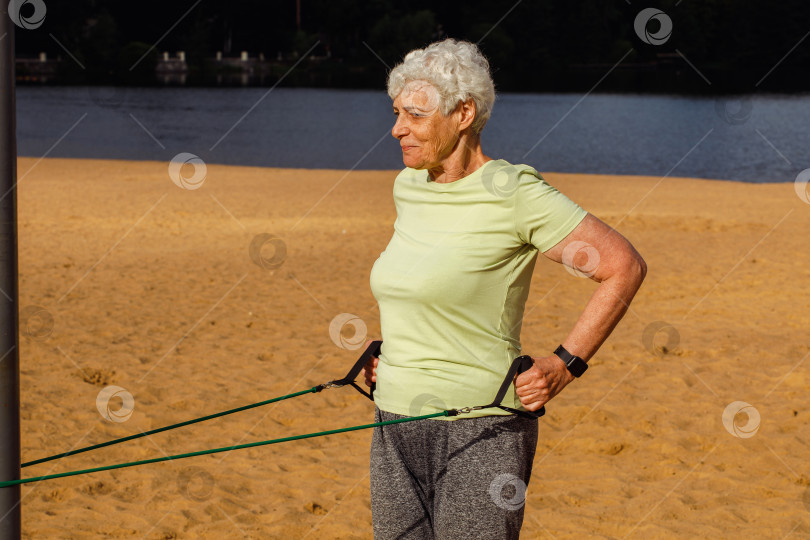 Скачать Пожилая женщина в спортивной одежде утром делает зарядку на открытом воздухе на пляже, используя эластичные резинки. фотосток Ozero