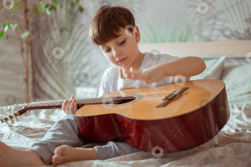 Скачать Маленький мальчик играет на гитаре дома, сидя на кровати в утреннем свете спальни фотосток Ozero
