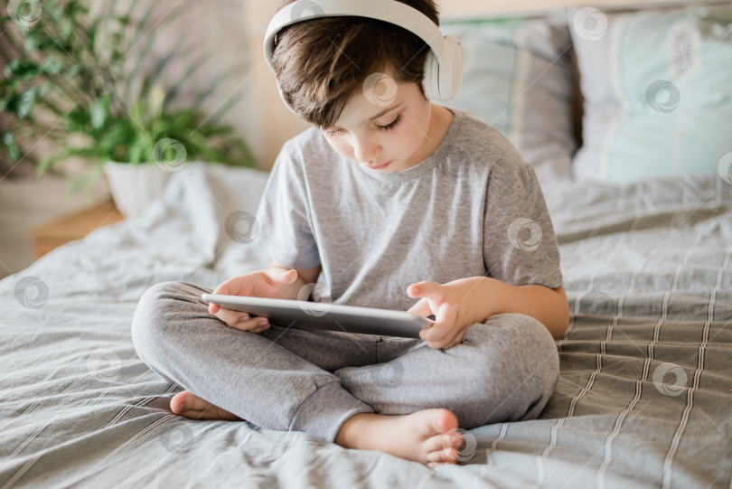 Скачать Счастливый ребенок лежит дома на кровати и играет с ПК планшетом или читает онлайн и слушает музыку фотосток Ozero