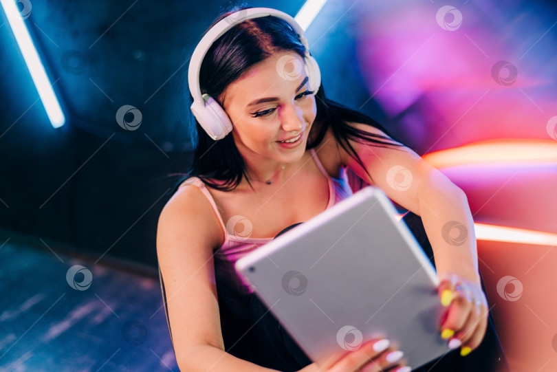 Скачать Современные устройства. Красивая молодая женщина, работающая на планшете с беспроводными наушниками в неоновом свете фотосток Ozero