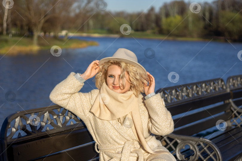 Скачать Женщина в бежевом пальто на берегу реки улыбается и придерживает шляпу от ветра на фоне весеннего пейзажа фотосток Ozero