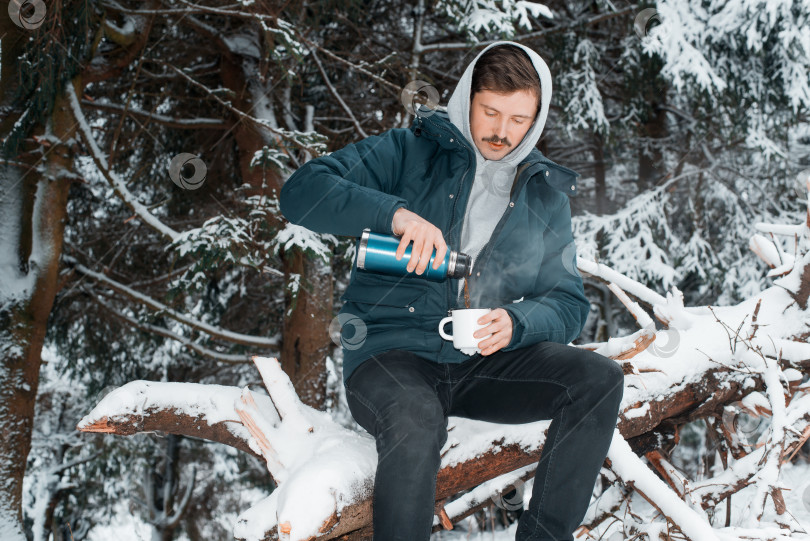 Скачать Путешественник разбивает лагерь в зимнем лесу. Мужчина наливает горячий напиток из термоса в кружку. Зимний заснеженный лес фотосток Ozero