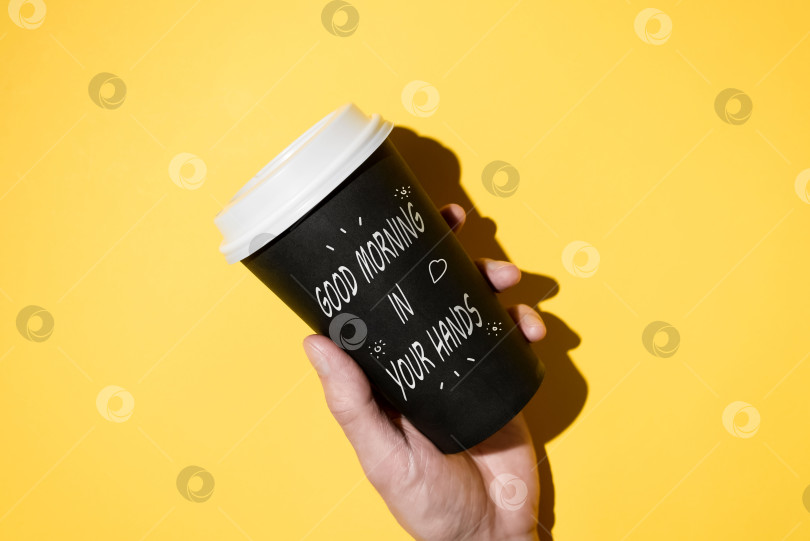 Скачать Концепция "Доброго утра". Рука, держащая одноразовую чашку кофе на желтом фоне. Надпись на картонном стаканчике "ДОБРОЕ УТРО В ВАШИХ РУКАХ" и нарисованные от руки сердечко и солнце на поверхности. фотосток Ozero