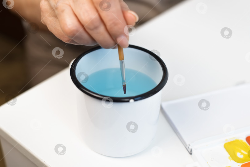 Скачать Женские руки отмывают кисть от краски в кружке с водой, крупный план. Избирательный фокус фотосток Ozero