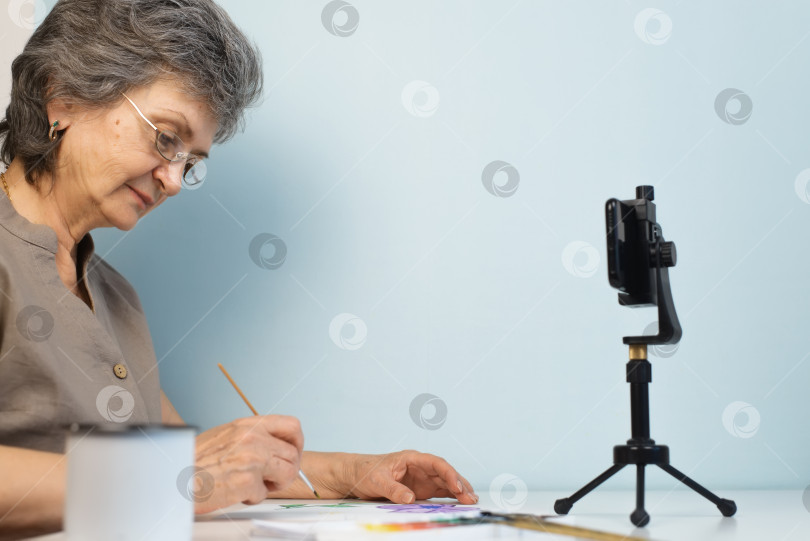 Скачать Онлайн-мастер-класс по рисованию. Пожилая женщина в очках рисует акварелью в помещении. Хобби на пенсии, концепция онлайн-курса. фотосток Ozero