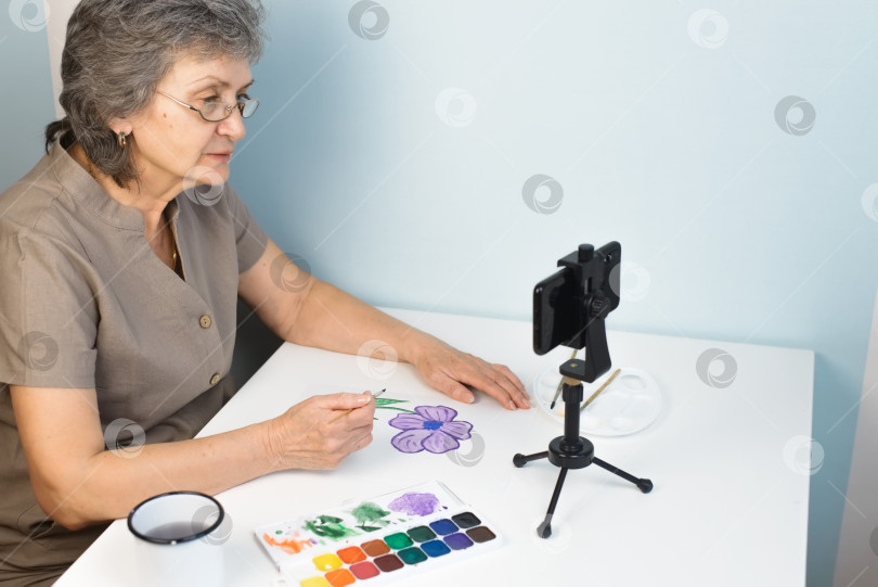 Скачать Женщина на пенсии смотрит онлайн-курс рисования. Пожилая женщина в очках рисует цветы акварелью в помещении. Дистанционное обучение на дому, концепция хобби на пенсии фотосток Ozero