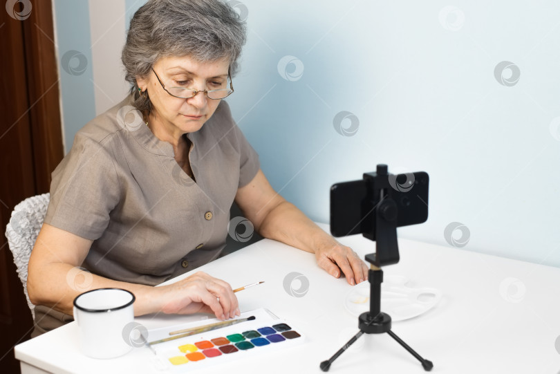 Скачать Пожилая женщина в очках рисует дома акварельную картину. Пенсионерка, проходящая онлайн-курс рисования. Концепция дистанционного обучения на дому фотосток Ozero