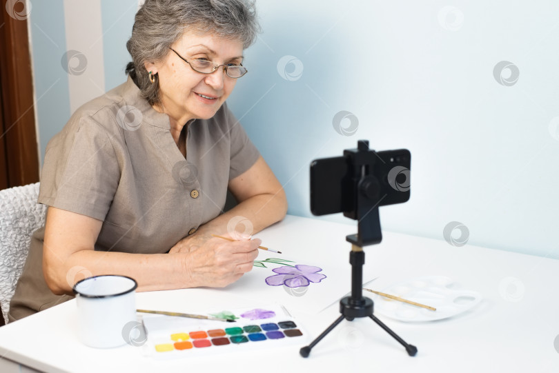 Скачать Пожилая женщина, проходящая онлайн-курс рисования. Пожилая женщина в очках рисует цветы акварелью в помещении. Дистанционное обучение на дому, концепция хобби на пенсии фотосток Ozero