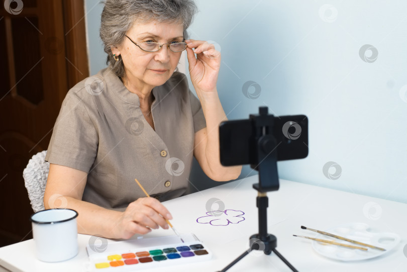 Скачать Пожилая женщина в очках смотрит на телефон. Онлайн-мастер-класс по рисованию. Пенсионерка рисует цветок акварелью в помещении. Хобби на пенсии, концепция онлайн-курса. фотосток Ozero