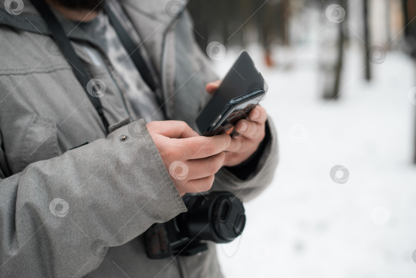 Скачать Фотограф пользуется мобильным телефоном на улице зимой. Мужчина в куртке и с фотоаппаратом, крупный план. Избирательный фокус фотосток Ozero