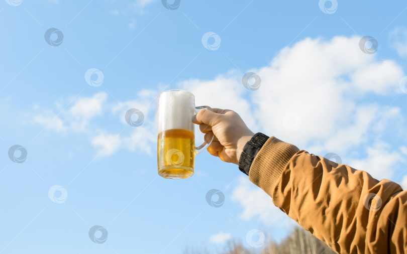 Скачать Мужская рука, держащая кружку пенистого пива на фоне голубого неба с облаками, на открытом воздухе. фотосток Ozero