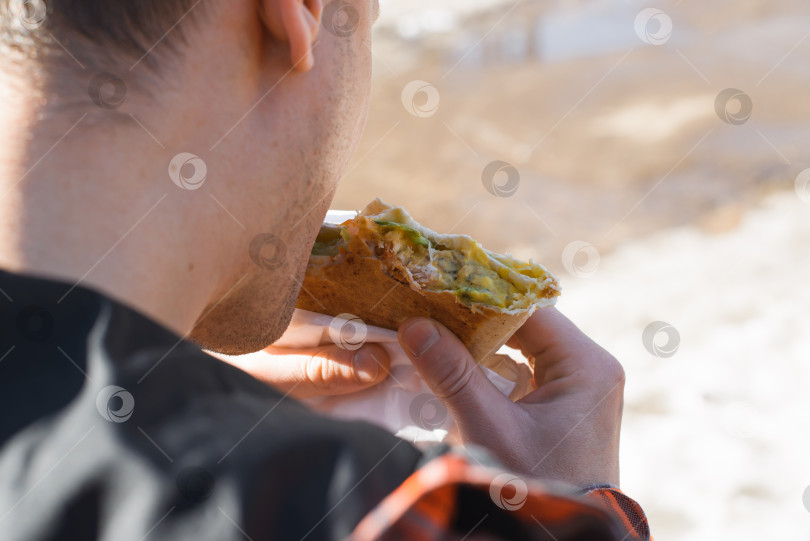 Скачать Мужчина ест фаст-фуд на улице в течение дня. Мужские руки, держащие острую уличную еду, крупным планом. Вид сзади фотосток Ozero