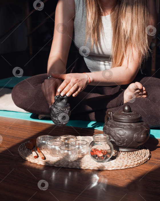 Скачать Чайная церемония в комнате для йоги и медитации на рассвете. Женщина, сидящая в позе лотоса, заваривает травяной чай, вид сверху. Глиняный чайник, деревянные ложки на подносе и поднимающийся пар фотосток Ozero