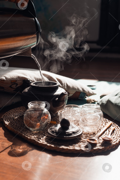 Скачать Женщина наливает горячую воду в глиняный чайник, заваривает травяной чай. От напитка, кружки и деревянных ложек на подносе поднимается пар. Чайная церемония на рассвете. фотосток Ozero