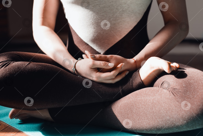 Скачать Молодая женщина в спортивном костюме практикует медитацию, сидя в позе лотоса на коврике для йоги, в помещении на рассвете. Крупный план. Концепция медитации осознанности, здорового образа жизни фотосток Ozero