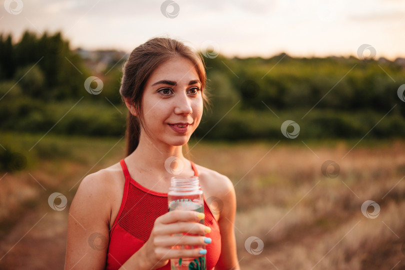 Скачать Красивая женщина-спортсменка по фитнесу пьет воду после тренировки, тренируясь летним вечером на закате в парке на открытом воздухе фотосток Ozero