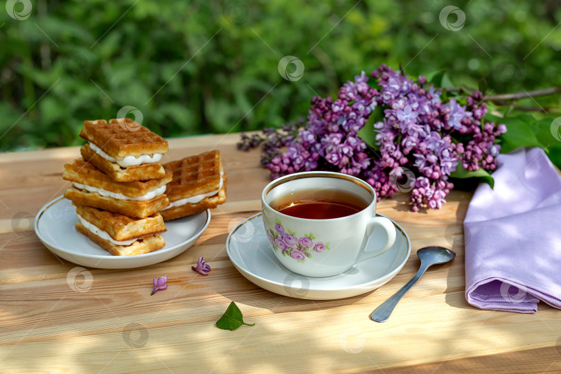 Скачать Чаепитие с венскими вафлями и веткой сирени на деревянном столе на фоне сада солнечным летним днем. фотосток Ozero