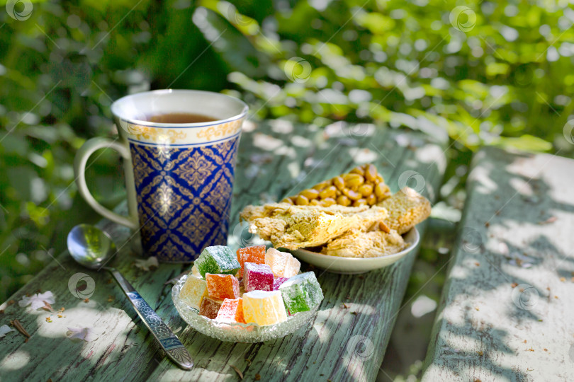 Скачать Восточные сладости и чай в чашке с рисунком среди зелени природы солнечным летним утром. Чаепитие в восточном стиле. фотосток Ozero