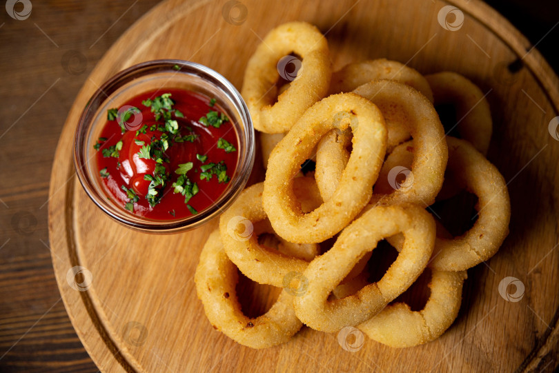 Скачать Луковые кольца с кетчупом выложите на деревянную доску. Еда в баре. Одна порция фотосток Ozero