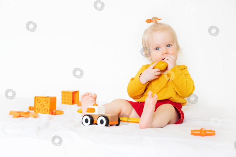Скачать Маленькая кавказская девочка играет с игрушками на белом фоне. Ребенку 1 год, он одет в оранжевую футболку и играет с оранжевыми игрушками. Фотография высокого качества фотосток Ozero