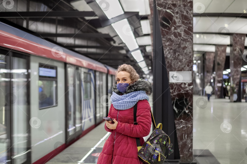 Скачать Молодая женщина в медицинской маске и ярко-пурпурном пальто с шарфом, держащая в руке телефон, ждет поезда в метро фотосток Ozero
