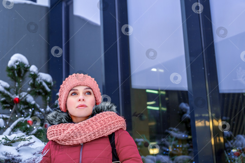 Скачать Молодая женщина в розовой шапочке и шарфе в заснеженном городе зимой у рождественской елки. Смотрит в будущее с надеждой. Крупный план. Место для текста фотосток Ozero