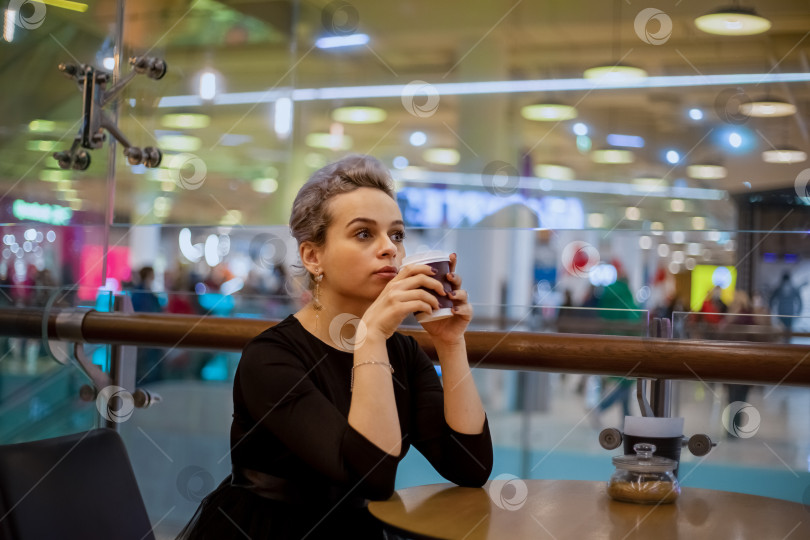 Скачать Молодая деловая женщина в черном платье пьет кофе в кафе, наслаждаясь одиночеством и думая о будущем. Отдых от работы фотосток Ozero