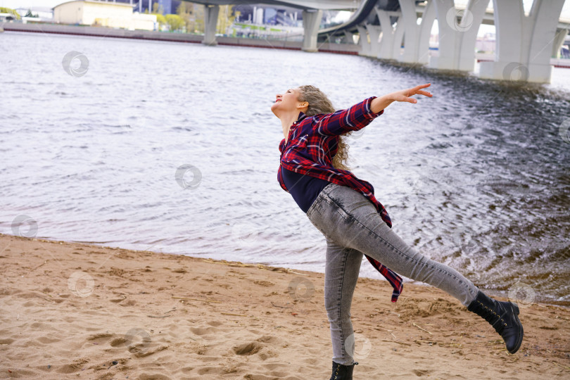 Скачать молодая красивая женщина в модной одежде позирует на фоне реки весенним солнечным днем, отдыхая с раскинутыми в стороны руками фотосток Ozero