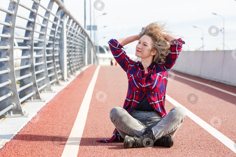 Скачать Усталая счастливая женщина отдыхает после бега, сидя на беговой дорожке и улыбаясь фотосток Ozero
