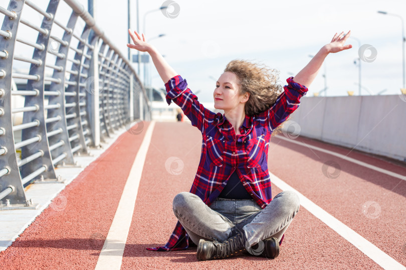 Скачать Уставшая счастливая женщина отдыхает после бега, сидя на беговой дорожке и улыбаясь фотосток Ozero