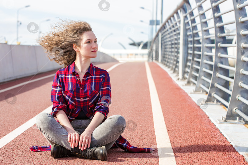 Скачать Усталая счастливая женщина отдыхает после бега, сидя на беговой дорожке и улыбаясь фотосток Ozero