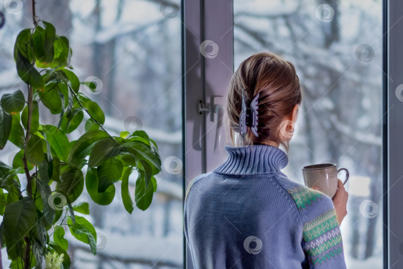 Скачать Женщина в синем свитере у окна со снежным пейзажем скучает и пьет кофе. Одиночество зимой фотосток Ozero