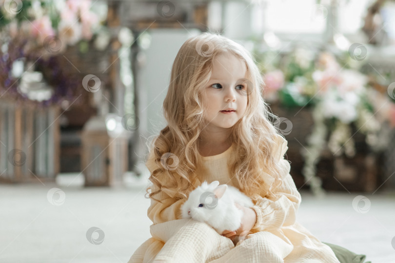Скачать Маленькая улыбающаяся девочка в желтом платье играет на полу в комнате с кроликами. Младенец и кролик. Гостиная украшена к Пасхе. фотосток Ozero
