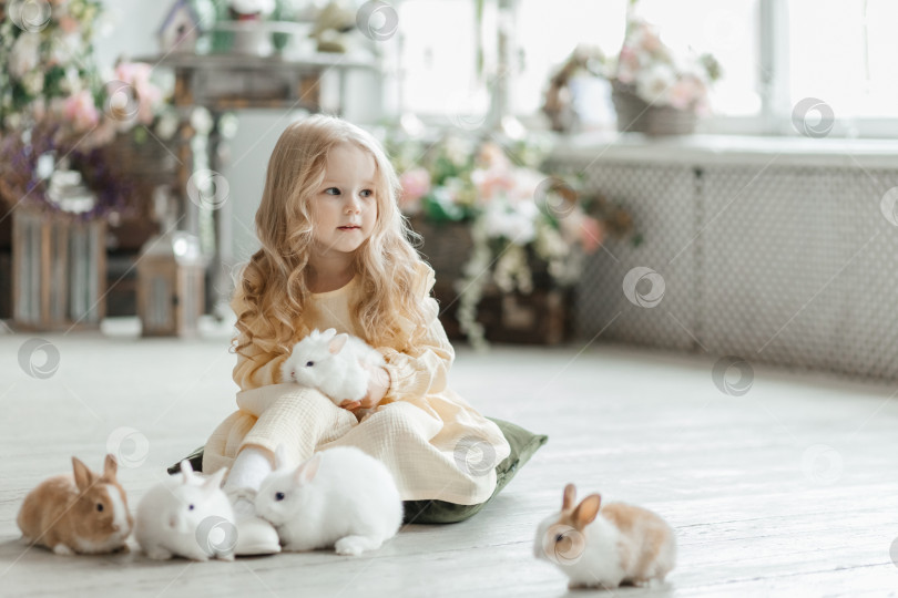 Скачать Маленькая улыбающаяся девочка в желтом платье играет на полу в комнате с кроликами. Младенец и кролик. Гостиная украшена к Пасхе. фотосток Ozero