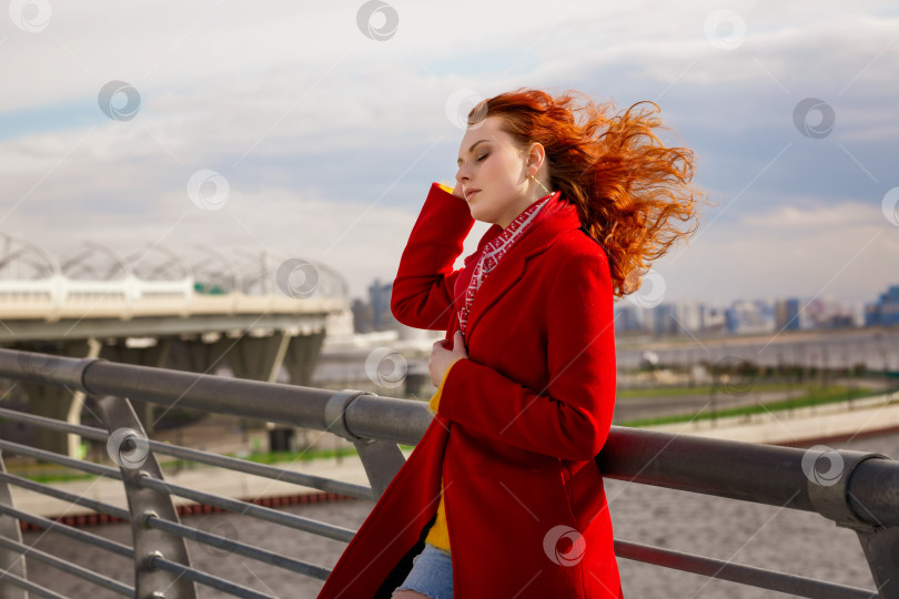 Скачать женщина в красном пальто стоит на мосту, поправляя волосы на ветру. фотосток Ozero