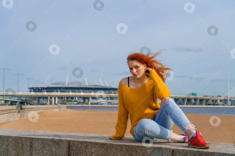Скачать Привлекательная молодая рыжеволосая женщина, сидящая снаружи на набережной, одетая в желтое фотосток Ozero