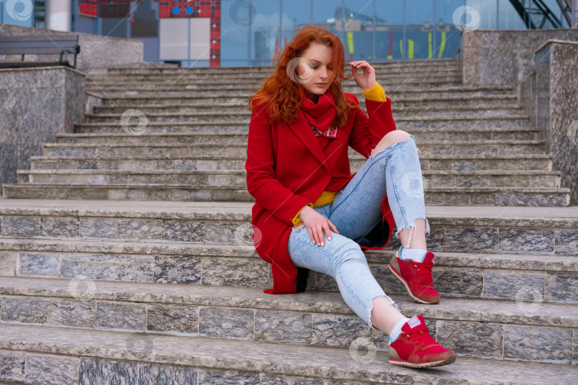 Скачать модная красивая девушка сидит на ступеньках возле офисного здания в красной куртке, джинсах и красных кроссовках фотосток Ozero