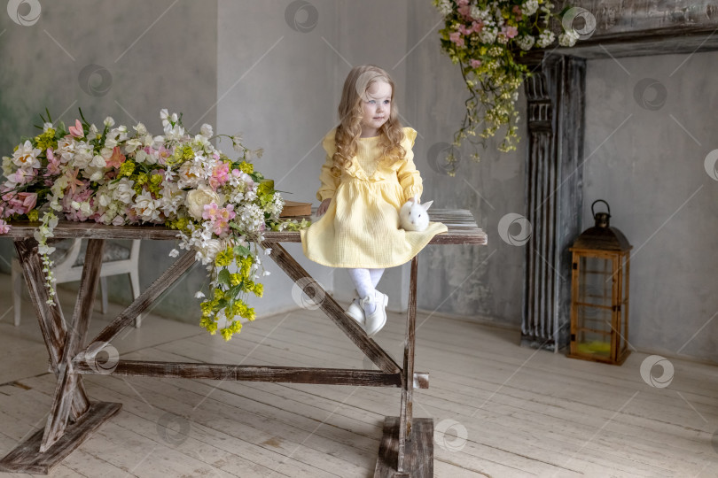 Скачать Маленькая улыбающаяся девочка в желтом платье сидит с кроликами на деревянном столе с цветами.Младенец и кролик. Концепция Пасхи. фотосток Ozero