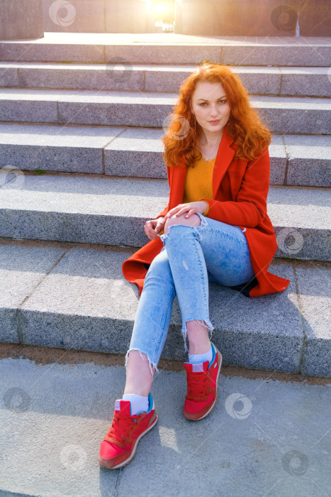Скачать Молодая рыжеволосая женщина в красном пальто, желтом свитере и джинсах сидит на лестнице фотосток Ozero