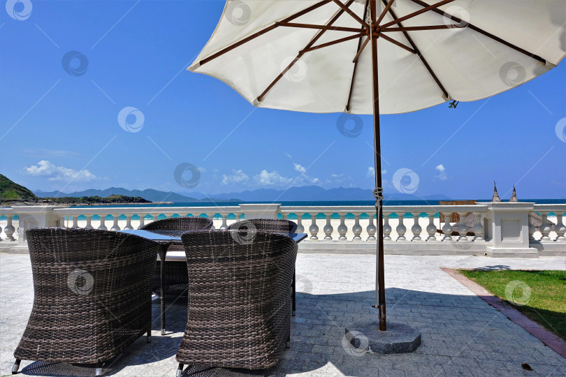 Скачать Отпуск во Вьетнаме. На набережной, в тени зонта, стоят стол и плетеные стулья. фотосток Ozero