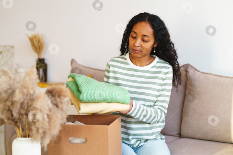 Скачать Афроамериканка упаковывает одежду для благотворительного пожертвования в картонную коробку в гостиной фотосток Ozero