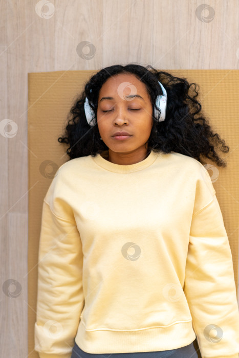 Скачать Афроамериканка в наушниках лежит на коврике с закрытыми глазами. Расслабление после тренировки фотосток Ozero