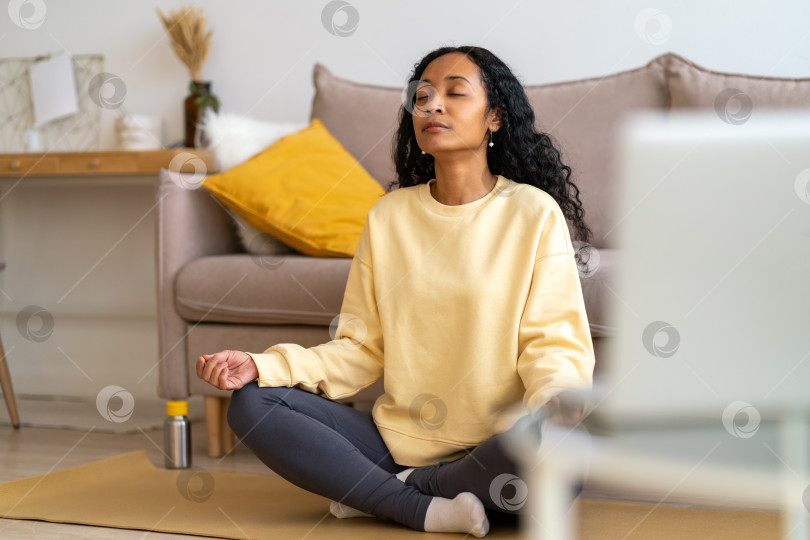 Скачать Молодая афроамериканка сидит в позе лотоса на коврике в гостиной во время медитации фотосток Ozero