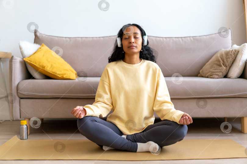 Скачать Молодая афроамериканка в наушниках слушает музыку во время медитации на коврике в гостиной фотосток Ozero