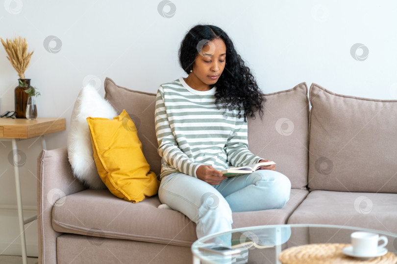 Скачать Афроамериканка читает книгу, сидя на диване в уютной гостиной. Проводить время в одиночестве фотосток Ozero