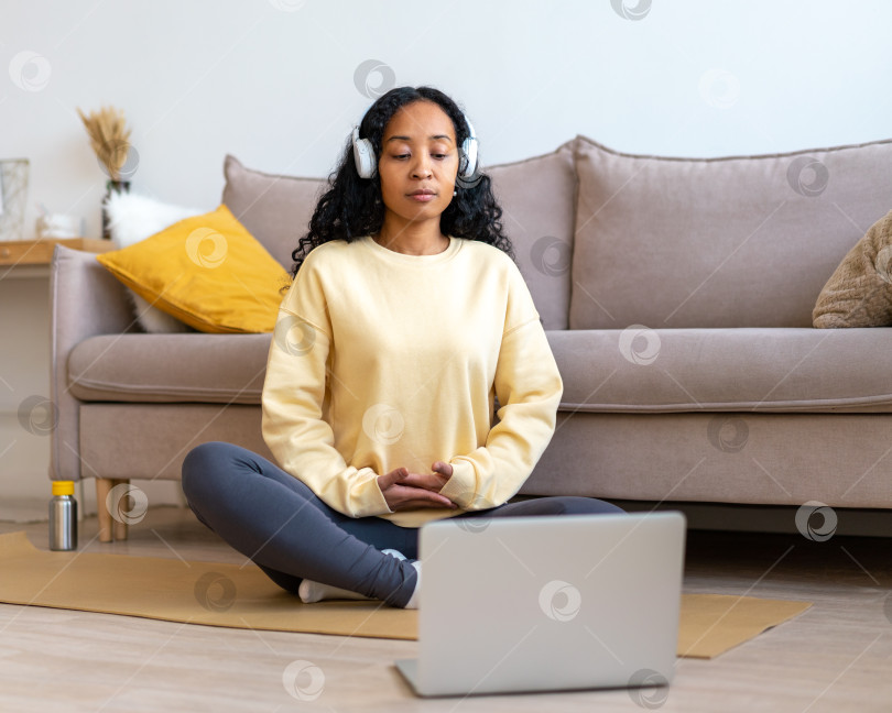 Скачать Афроамериканка слушает музыку в наушниках во время медитации с ноутбуком дома фотосток Ozero