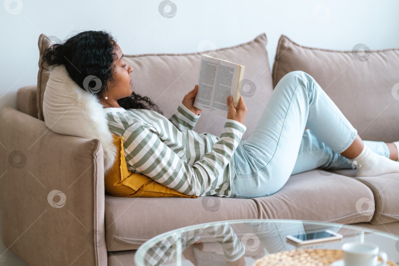 Скачать Афроамериканка отдыхает на диване и читает книгу. Отключение от сети для цифровой детоксикации фотосток Ozero