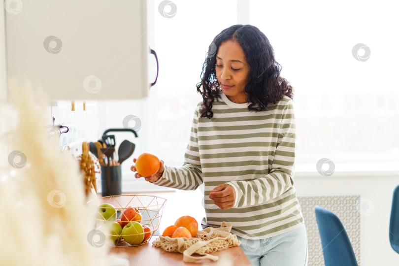 Скачать Афроамериканка на кухне перебирает яблоки и апельсины после покупок в продуктовом магазине фотосток Ozero